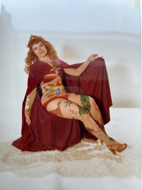 A middle-aged woman poses with a cape, displaying tattoos covering her body.